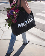 Image of a woman walking across the street with the MUTHA Tote Bag filled with flowers on her shoulder