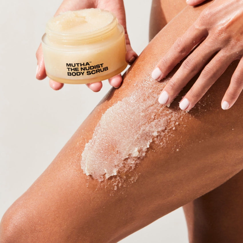 Image of a model applying a swipe of the NUDIST Body Scrub to the thigh while holding the jar in the other hand