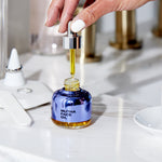 Image of the Face Oil on a filled marble countertop with a hand holding up the dropper of the Face Oil